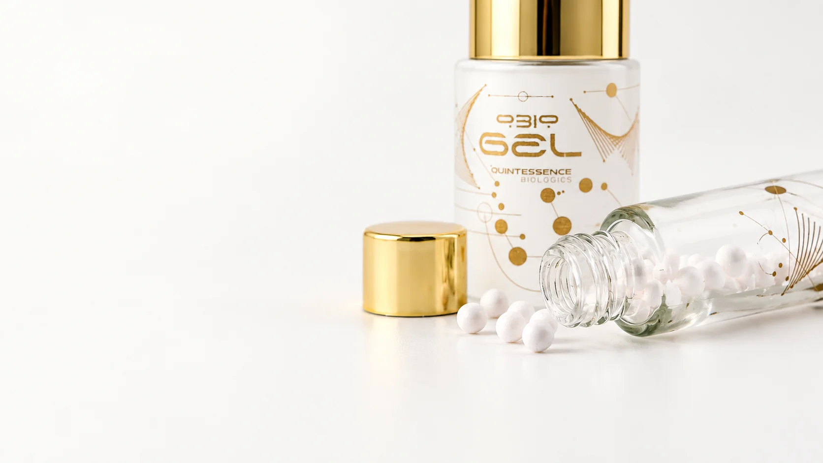 An open QBio Elixir vial with white lyophilized beads, its gold cap, and QBio Gel on a pale studio surface.